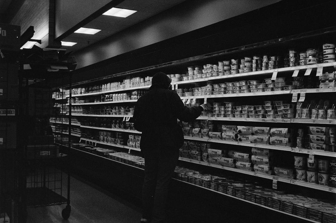 Pessoa de costas em corredor de supermercado observando prateleiras refrigeradas cheias de potes de laticínios.